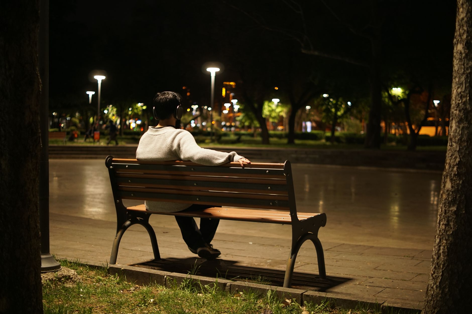 Solitary figure listening to music on a park bench under night streetlights, urban solitude.
