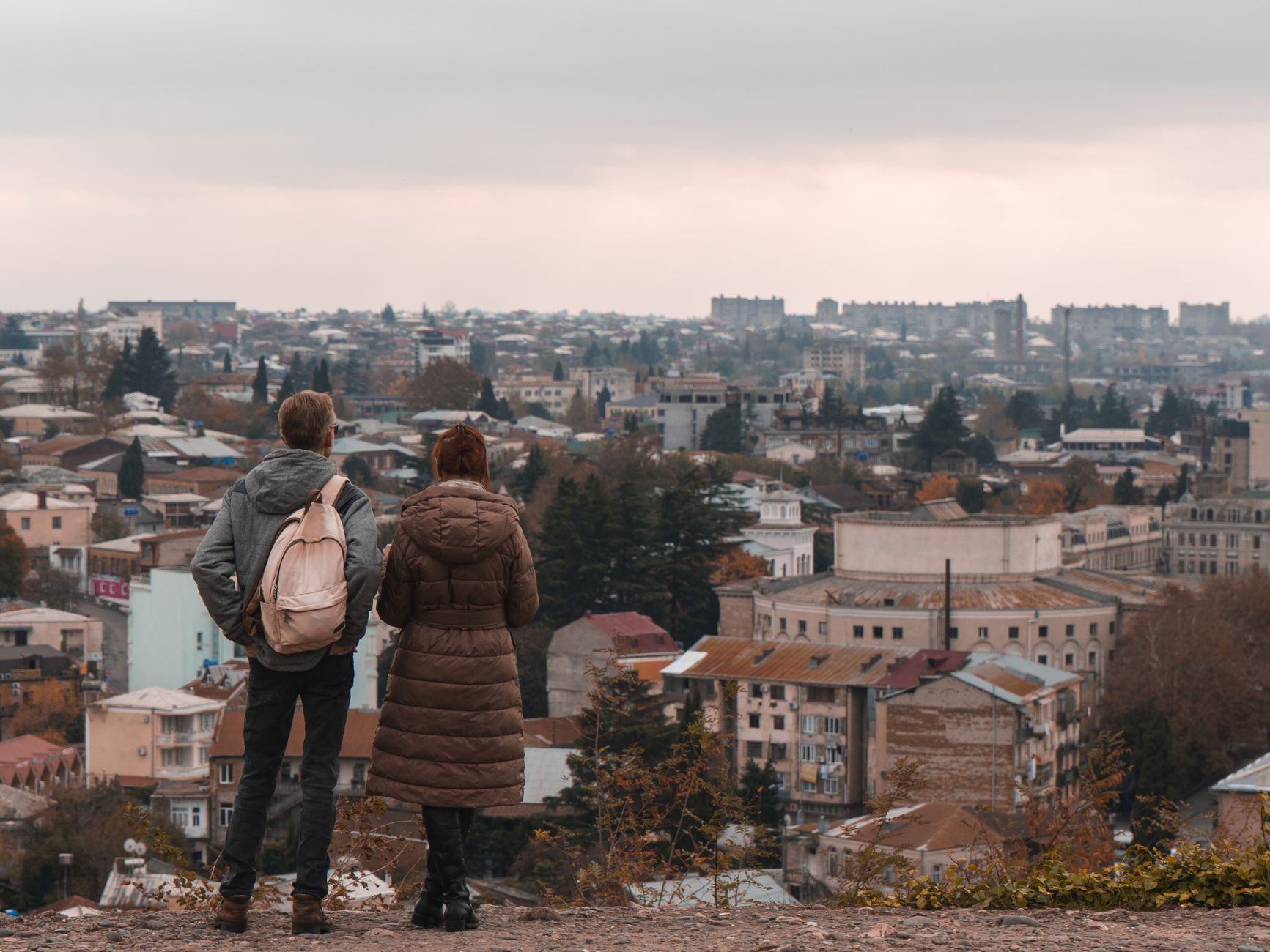A couple stands on a hill, overlooking a vast townscape with autumn foliage under a cloudy sky.