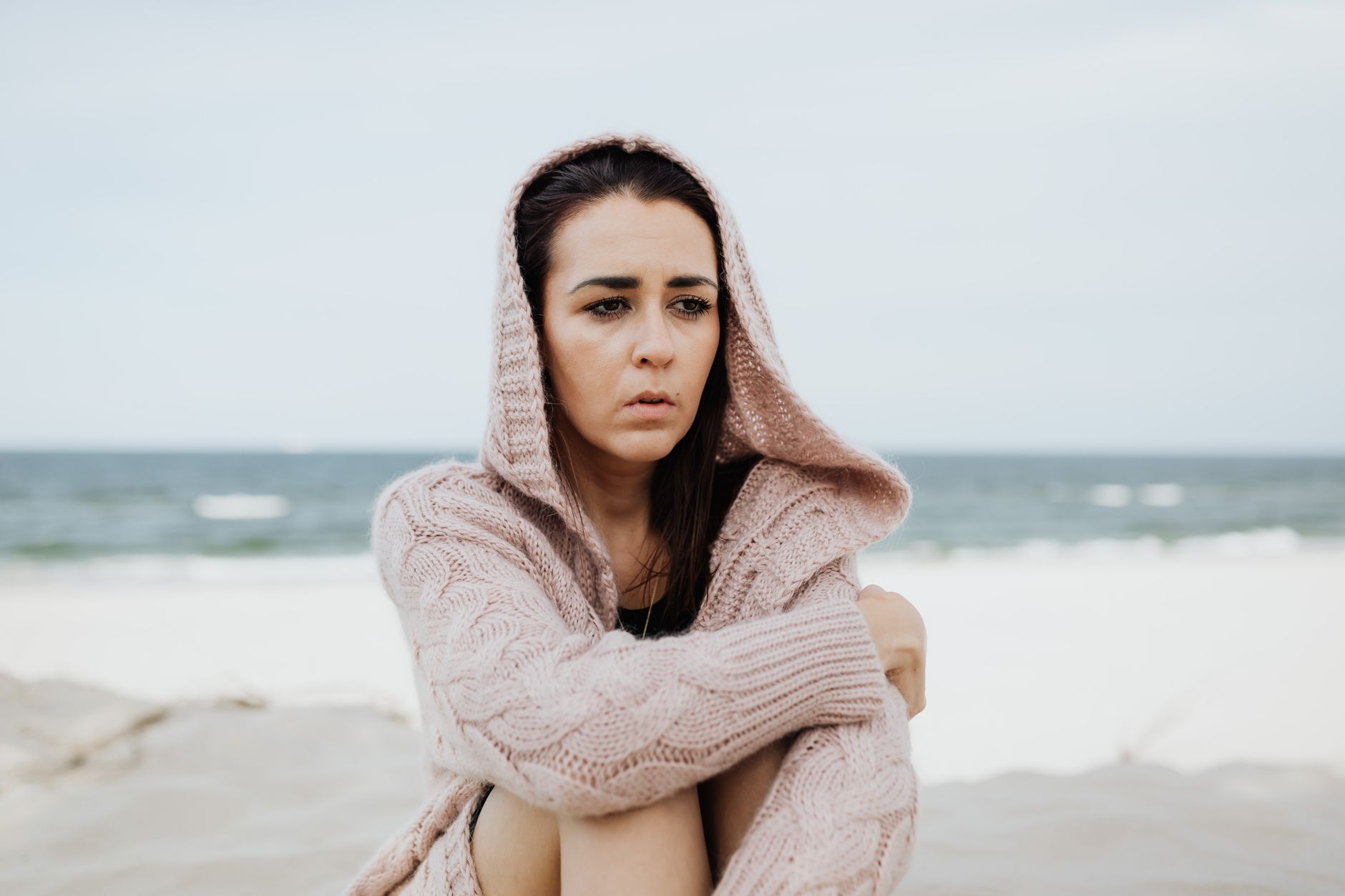A woman in a hooded sweater sits pensively on a windy beach, gazing into the distance.