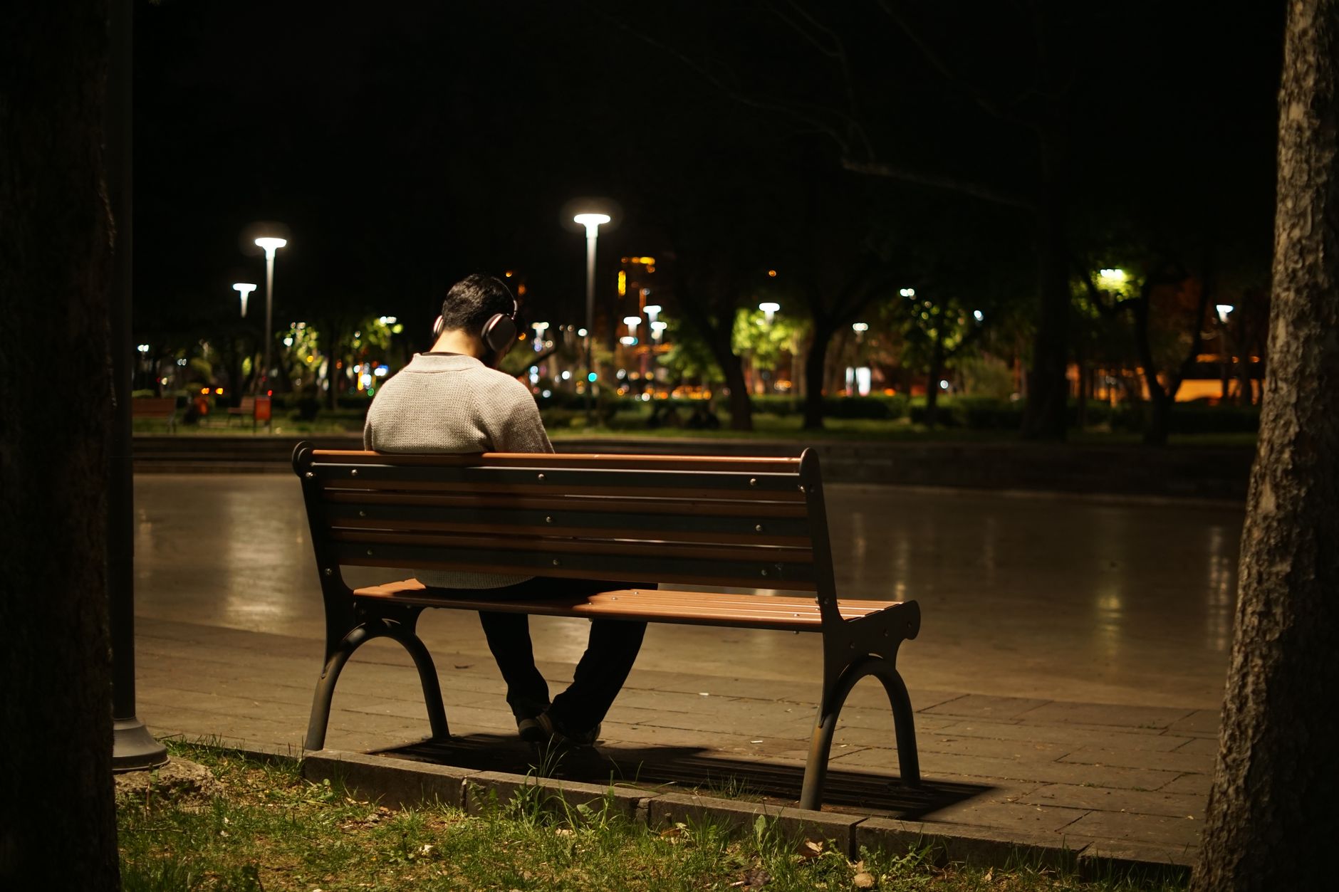 A man wearing headphones sits on a park bench at night, depicting solitude and urban loneliness.