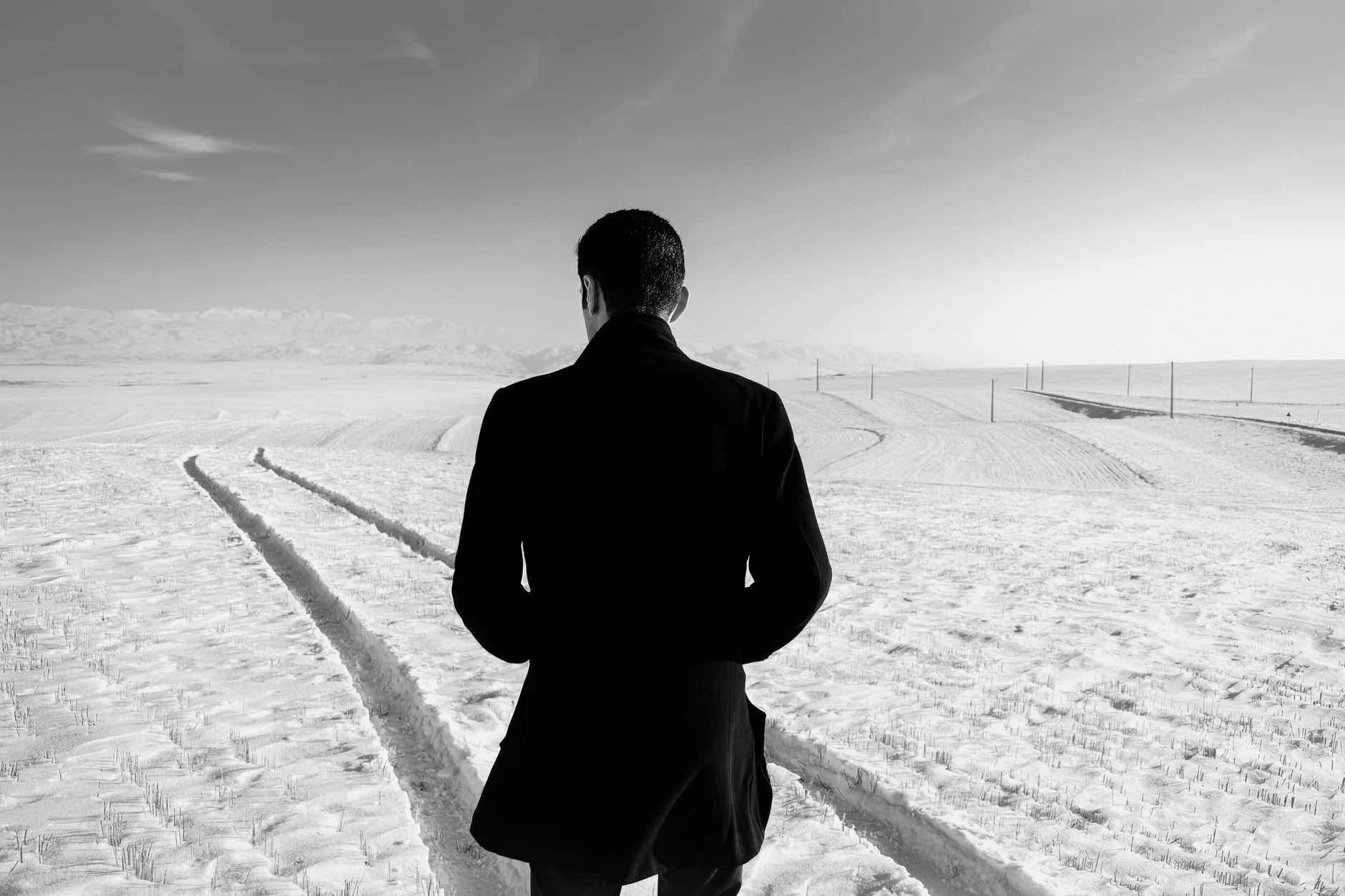 Black and white back view of anonymous male wearing warm coat with hands on pockets standing in middle of snowy terrain