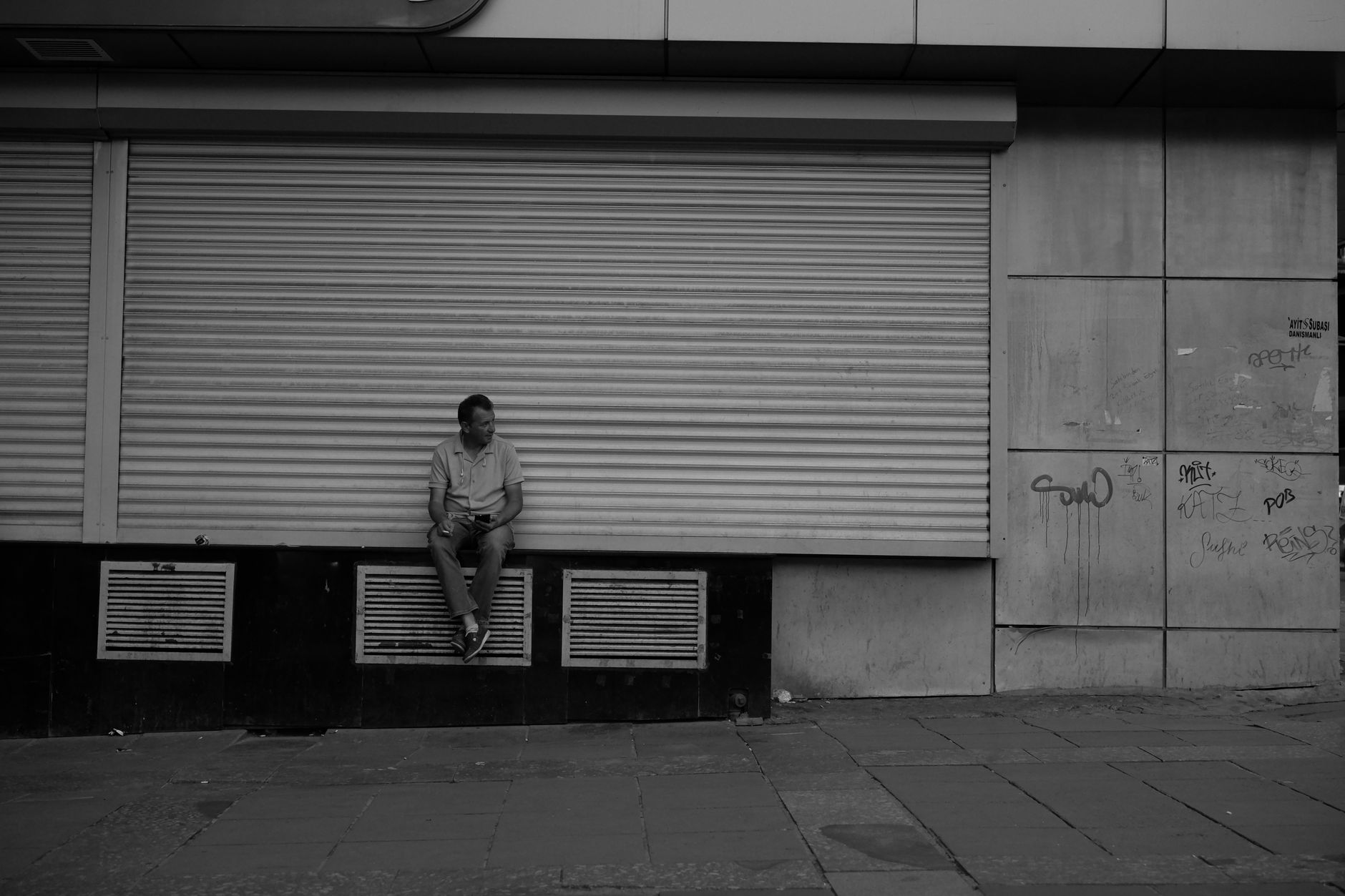 Black and white photo of a man sitting outside a closed store in an urban setting.