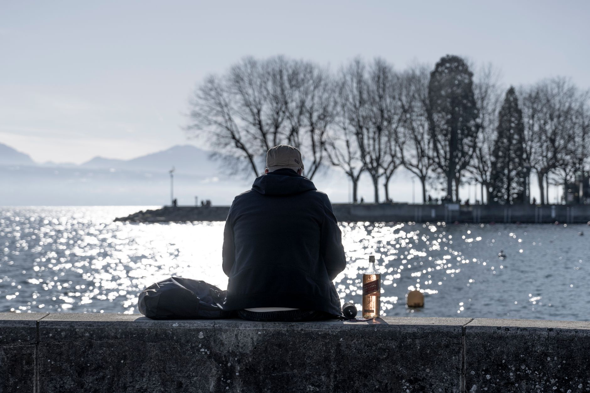 A solitary man in a jacket facing a body of water in a park, reflecting on life.
