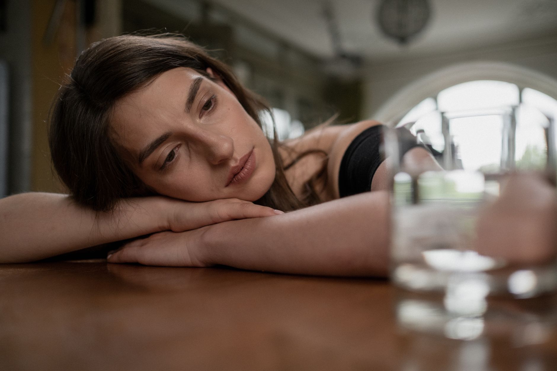 Pensive woman resting her head on a wooden table with a glass of water nearby, indoors.