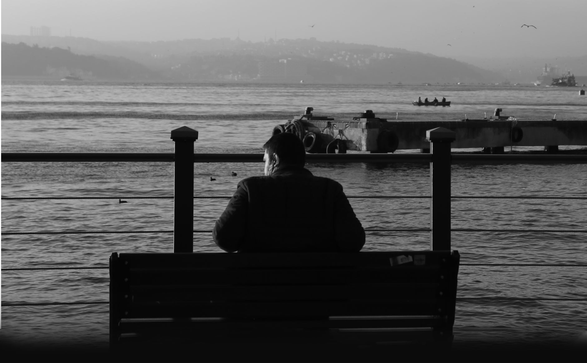 A man sits on a bench overlooking a tranquil body of water in a monochrome landscape.