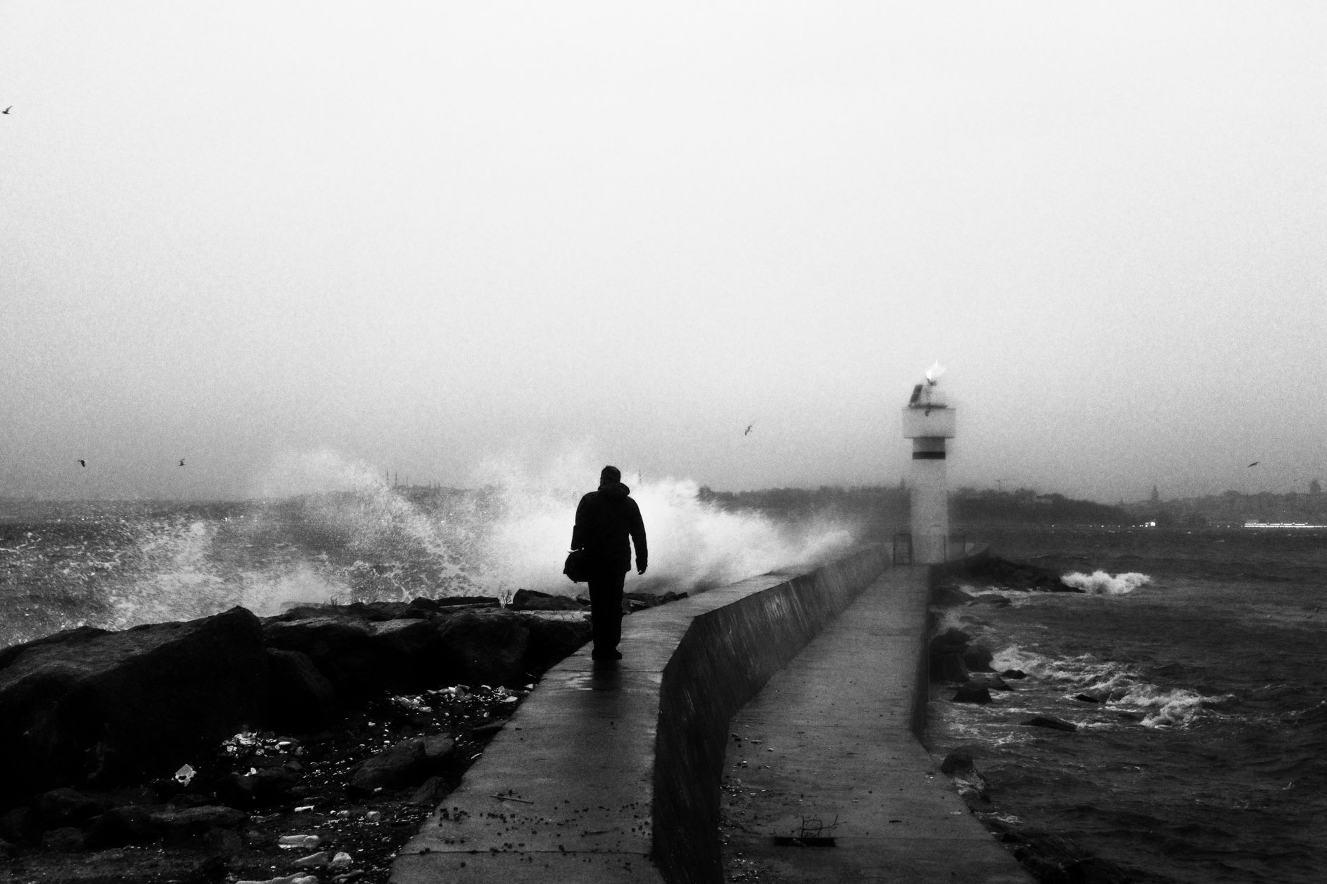 A lone silhouette walking on a breakwater with crashing waves and lighthouse in monochrome.